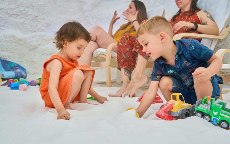 Toddlers playing with toy trucks in a salt cave during halotherapy session with mothers in the background